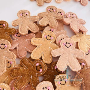 以下が含まれることがあります： ピンクの笑顔の顔をした、さまざまな茶色のフェルトで作られたジンジャーブレッドマンのオーナメントのコレクション。オーナメントはクリスマスツリーの飾り付けや、ホリデーデコレーションにフェスティブなタッチを加えるのに最適です。