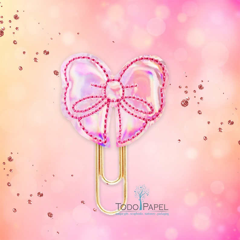 May include: A gold paper clip with a pink iridescent bow attached. The bow has a pink stitched outline. The text "TODO PAPEL" is visible in the bottom right corner of the image.