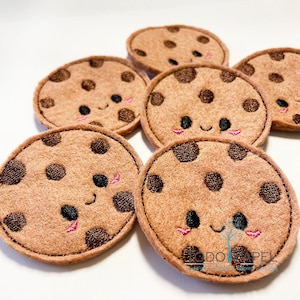 Op de afbeelding: Zes bruine vilt chocoladekoekjes met geborduurde gezichten en chocolade chips. De koekjes hebben roze wangen en zwarte ogen.