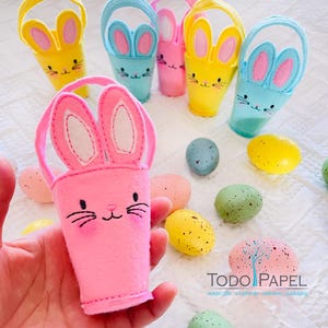 May include: A set of felt bunny baskets in pink, yellow, and blue. The baskets are decorated with embroidered bunny faces and ears. The baskets are filled with colorful speckled Easter eggs.