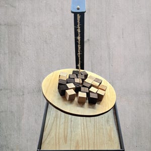 Peut inclure: Un support vertical en métal noir soutient une base rectangulaire en bois et une plateforme circulaire suspendue. Une ficelle pend du bras supérieur, retenant la plateforme sur laquelle sont disposés de petits cubes en bois clairs et foncés.