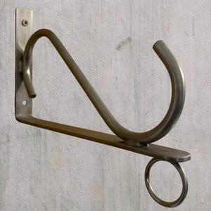 Könnte beinhalten: Ein silberfarbener Wandhalter aus Metall mit einer flachen, vertikalen Montageplatte mit zwei Schraublöchern, einem horizontalen Stützarm, einer geschwungenen dekorativen Strebe und einem kleinen runden Ring an der Unterseite des Arms.
