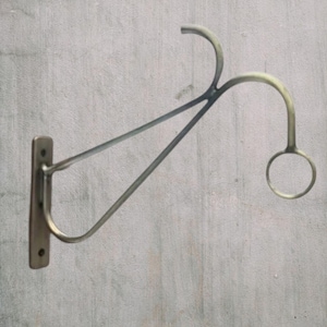 Op de afbeelding: Een wandbeugel van metaal met een rechthoekige basisplaat met twee schroefgaten, een gebogen steunarm en een cirkelvormige ring aan het uiteinde. Het metaal heeft een matte, geborstelde afwerking met subtiele gouden en grijze tinten.