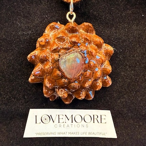 Puede incluir: Colgante artesanal con diseño de piña marrón texturizada y una piedra iridiscente central. Cuelga de un cordón de cuero marrón. La etiqueta dice "LOVE MOORE CREATIONS" y "PRESERVING WHAT MAKES LIFE BEAUTIFUL".