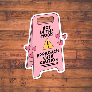 May include: A pink sticker in the shape of a caution sign with the words "NOT IN THE MOOD" and "APPROACH WITH CAUTION" in black. A yellow triangle with an exclamation point is in the center. Pink hearts are scattered around the sign.