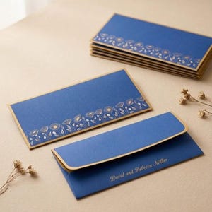 May include: A set of rectangular blue envelopes with gold-colored edges. One envelope is open, displaying a gold-colored floral line art pattern along the bottom. Another envelope is closed, featuring the gold text David and Rebecca Miller.