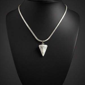 Op de afbeelding: Een zilveren ketting met een witte, driehoekige hanger. De hanger heeft een gemarmerd, zwart-wit patroon. De ketting wordt gepresenteerd op een zwarte mannequin. De ketting heeft een dun, geweven ontwerp.