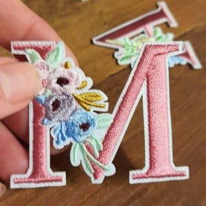 Può includere: Toppa ricamata con la lettera "M" rosa con accenti floreali nelle tonalità del rosa, blu e viola. La toppa ha un bordo bianco. Un'altra toppa con lettera è visibile sullo sfondo.