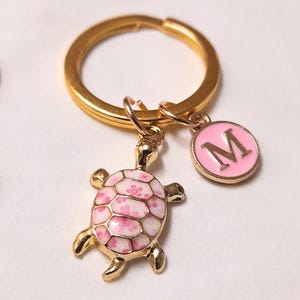 Peut inclure: Un porte-clés doré avec un charm en forme de tortue et un charm rose avec la lettre "M". Le charm de la tortue a un motif de carapace rose et blanc. Accessoire décoratif.