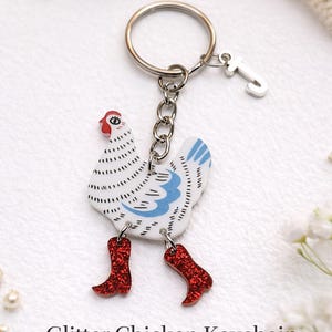 May include: A whimsical chicken keychain with a white body, blue wing accents, and red glitter boots. The keychain includes a silver keyring and a small charm. The text "Glitter Chicken Keychain" and "Funny Farm Animal Key Ring" are visible.