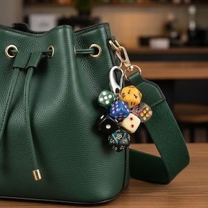 Può includere: Una borsa a secchiello in pelle verde scuro con un portachiavi a dadi. Il portachiavi ha diversi dadi colorati attaccati a un fermaglio argentato. La borsa ha finiture color oro e una tracolla verde.