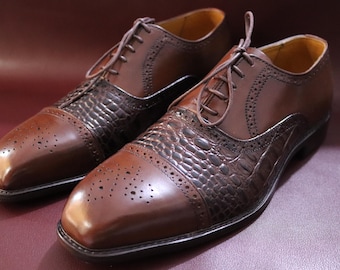 Puede incluir: Un par de zapatos de vestir de cuero marrón con un patrón texturizado similar al de un caimán en la puntera y la pala. Los zapatos tienen un diseño clásico con cordones y se exhiben sobre una superficie burdeos.