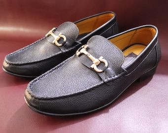 May include: A pair of black leather loafers with gold-colored metal accents. The shoes have a classic design with a rounded toe and a slip-on style. The loafers are displayed on a burgundy surface.