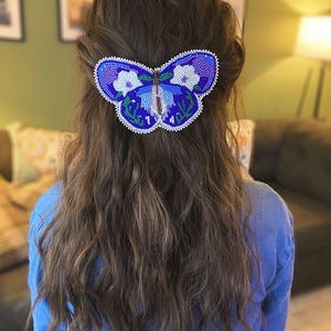 May include: A beaded butterfly hair clip in shades of blue, purple, and white. The butterfly design is intricately detailed with small beads, and is attached to a hair clip. The hair accessory is worn in the hair.