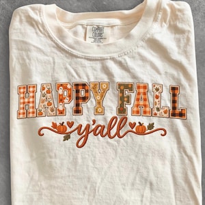 以下が含まれることがあります： アイボリー色の半袖Tシャツ。チェック柄、カボチャ、葉などの秋らしい模様が施されたブロック体の「HAPPY FALL」という文字がプリントされています。その下には、カボチャを添えた筆記体で「y'all」という言葉が書かれています。