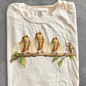 Op de afbeelding: Een ivoorkleurig t-shirt met een grafische print van drie cicaden en één sprinkhaan op een bruine boomtak met groene bladeren. De cicaden hebben doorschijnende vleugels en bruine lichamen, terwijl de sprinkhaan kleiner is en een bruine kleur heeft.