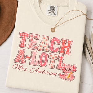 May include: An ivory t-shirt featuring the text TEACH A-LOTL and Mrs. Anderson in red script. The letters have various red and pink patterns including gingham, floral, and polka dots. A cartoon axolotl wearing glasses and holding a pencil is included.