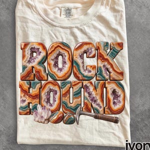 以下が含まれることがあります： アイボリーのTシャツ。オレンジ、ティール、紫の同心円状の帯を持つアゲートやアメジストのジオードのようなブロック体の「ROCK HOUND」という文字がプリントされている。文字の下にはアメジストの結晶、2つの滑らかな石、ハンマーが描かれている。