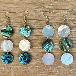 May include: Four pairs of dangle earrings with iridescent abalone shell and white shell discs. The earrings have gold and silver-colored hooks. The abalone shells have shades of green, blue, and teal.