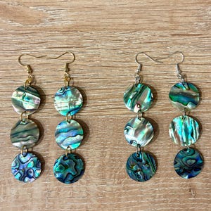 Könnte beinhalten: Vier Paar Ohrhänger mit schillernden Abalone-Muschelscheiben. Jedes Ohrring hat drei kreisförmige Muschelteile in Blau-, Grün- und Silbertönen. Die Ohrringe haben goldene und silberne Haken.