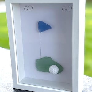 May include: A framed golf-themed art piece. A blue glass triangle represents a flag, connected by a thin wire to a green glass shape. A white golf ball rests on the green glass. The frame is white.