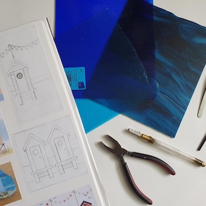 May include: An assortment of craft supplies, including blue stained glass sheets, a sketchbook with beach hut illustrations, pliers, tweezers, and a glass cutter. The tools and materials are arranged on a white surface, suggesting a crafting project.