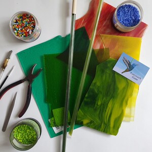 May include: A collection of stained glass supplies, including sheets of green, yellow, and red glass, glass beads, glass rods, and tools like pliers and a glass cutter. A business card with a hummingbird and the text "Fusionark Glass" is also included.