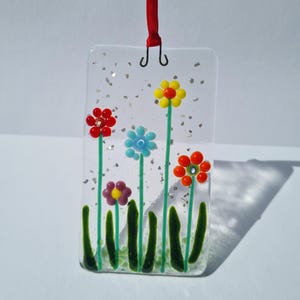 Puede incluir: Un adorno colgante rectangular de vidrio fundido con fondo transparente y motas plateadas. El diseño muestra cinco flores coloridas con tallos y hojas verdes, suspendido por una cinta roja sujeta a una anilla metálica en la parte superior.