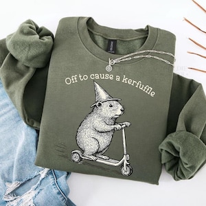 Puede incluir: Sudadera verde oliva con la frase "Off to cause a kerfuffle". La ilustración muestra un roedor con un sombrero de bruja montado en un patinete. Una cadena de plata está colocada sobre la sudadera.