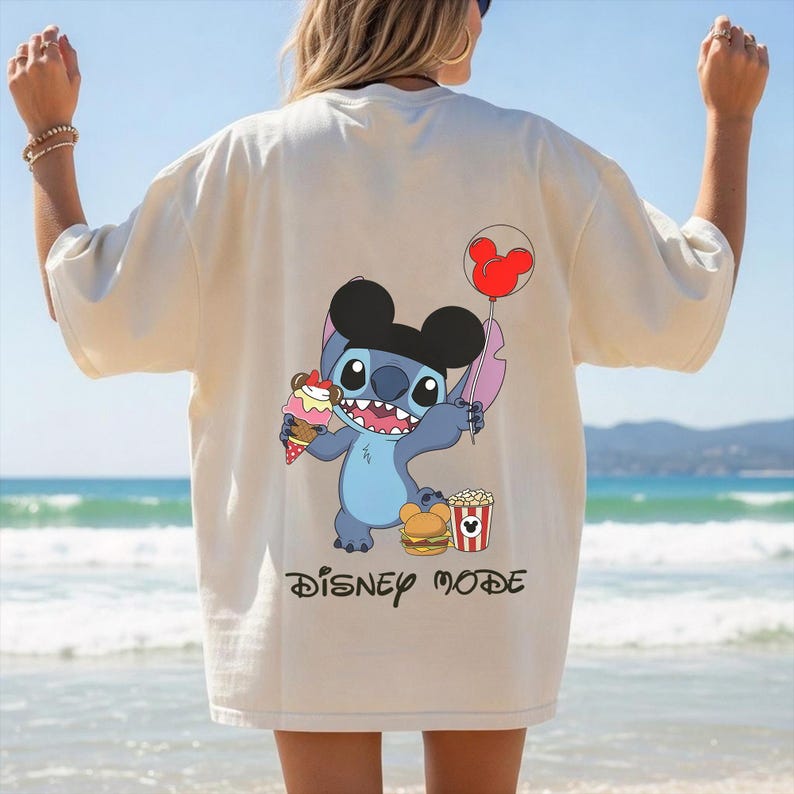 Peut inclure: T-shirt cr&egrave;me avec un personnage de dessin anim&eacute; Stitch portant un chapeau d'oreille de Mickey Mouse, tenant une glace et un ballon. Le t-shirt a &eacute;galement un burger et un pop-corn graphiques, avec les mots "DISNEY MODE" imprim&eacute;s en dessous.