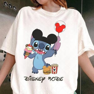 Peut inclure: T-shirt blanc cass&eacute; avec un personnage de dessin anim&eacute; bleu tenant un ballon Mickey Mouse rouge et un cornet de glace. Le personnage porte un chapeau noir. Le t-shirt porte l'inscription "DISNEY MODE" en noir.