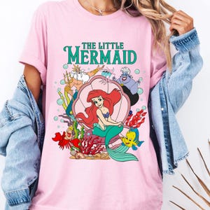 May include: Pink t-shirt featuring a colorful graphic of Ariel from The Little Mermaid, seated in a seashell with other characters. The words "THE LITTLE MERMAID" are printed above the image in green. The shirt has short sleeves.