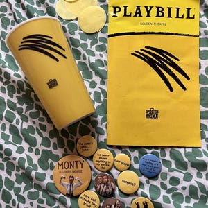Può includere: Un bicchiere giallo e un programma Playbill abbinato per Operation Mincemeat, entrambi con una grafica a forma di graffio nero. Sono incluse diverse spille rotonde con vari testi e disegni fotografici, oltre a quattro piccoli cerchi di carta gialla.
