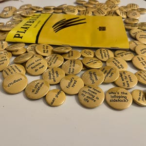 Könnte beinhalten: Eine Ansammlung beigefarbener Ansteckbuttons mit schwarzen Texten und ein gelbes Playbill. Die Buttons tragen Sätze wie "Wer ist der tollpatschige Sidekick?" und "Ich habe hier sechs Jahre gearbeitet".
