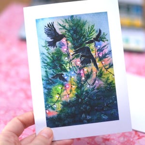 Könnte beinhalten: Ein Aquarelldruck mit einem Weihnachtsbaum und schwarzen Vögeln im Flug. Der Baum ist mit bunten Lichtern geschmückt, vor einem blau-grünen Hintergrund. Der Druck wird in der Hand einer Person gehalten.