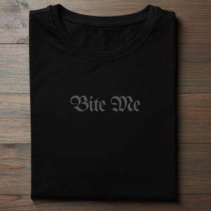 Folded black gothic typography shirt with charcoal Bite Me text minimalist alternative clothing design
