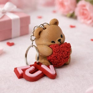 May include: A brown teddy bear keychain holding a red heart, with the word "LOVE" in red and pink letters. The background includes a pink gift box, red heart-shaped confetti, and pink roses.