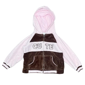 Może przedstawiać: Różowa i brązowa bluza z kapturem zapinana na zamek z napisem "CUTE" na piersi. Bluza ma jasnoróżowe wnętrze i przednią kieszeń. Rękawy i dolny brzeg mają brązowo-białe paski.