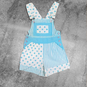 Op de afbeelding: Lichtblauwe en witte baby-overalls met een mix van stippen en verticale strepen. De slab heeft een vierkante zak met stippen en de bandjes zijn ook gestippeld. De shorts hebben knoopsluitingen aan de zijkanten.