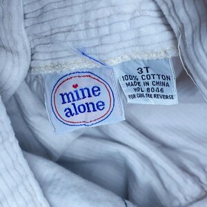 May include: Close-up of a white corduroy garment with a tag that reads "mine alone" in a red and blue circle. The tag also indicates the size is 3T and made of 100% cotton.
