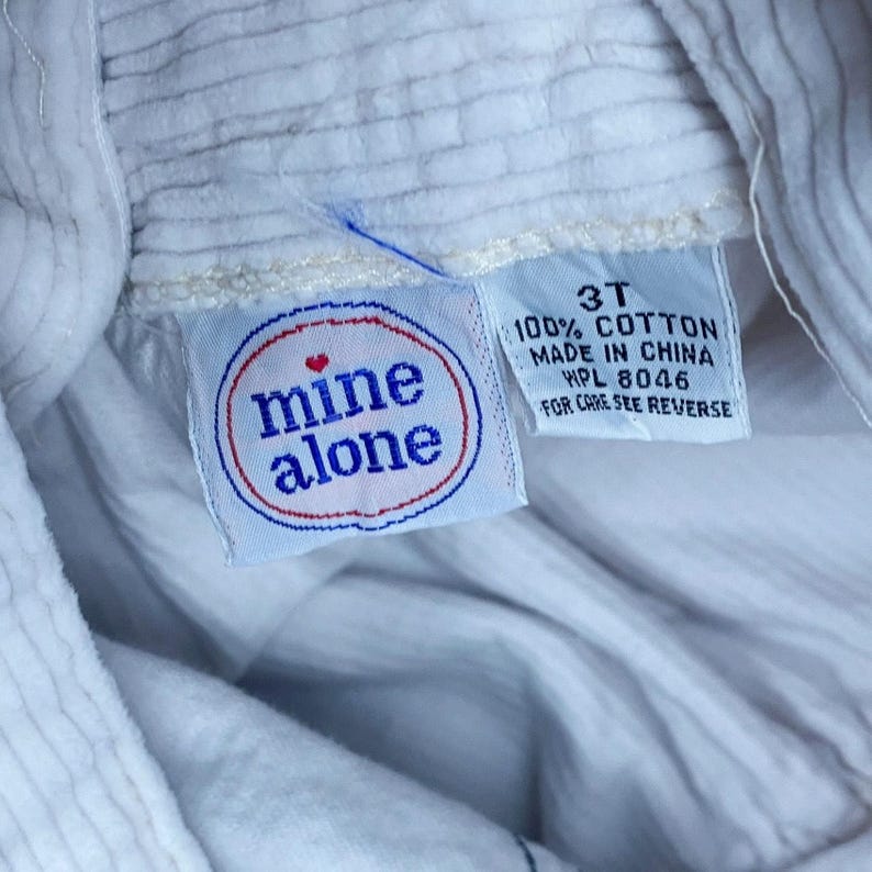 May include: Close-up of a white corduroy garment with a tag that reads "mine alone" in a red and blue circle. The tag also indicates the size is 3T and made of 100% cotton.