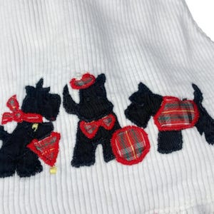 May include: White corduroy fabric with three black terrier dog appliqu&eacute;s. Each dog has red and plaid accents, including bows, hats, and a plaid ball. The dogs are embroidered with black thread.