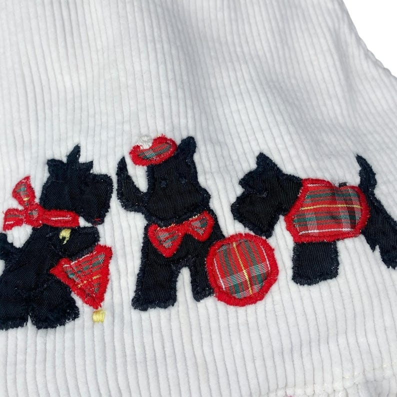 May include: White corduroy fabric with three black terrier dog appliqu&eacute;s. Each dog has red and plaid accents, including bows, hats, and a plaid ball. The dogs are embroidered with black thread.
