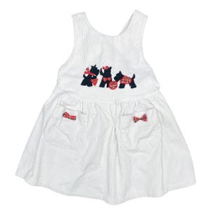 May include: White corduroy dress with a sleeveless design. Features three embroidered black dogs with red plaid accents and bows. Two pockets with red plaid bows are on the skirt. A classic, vintage style.