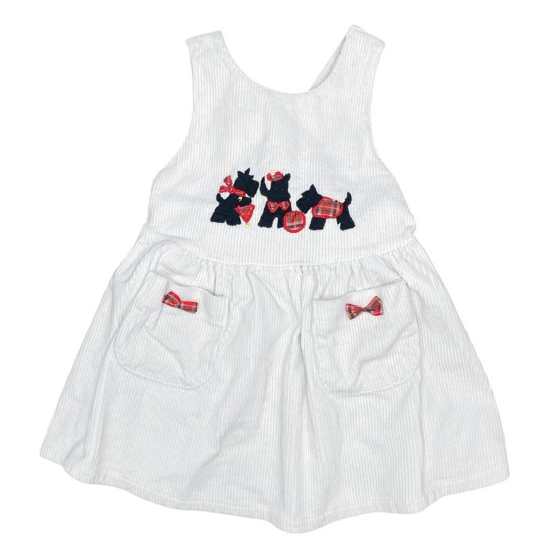 May include: White corduroy dress with a sleeveless design. Features three embroidered black dogs with red plaid accents and bows. Two pockets with red plaid bows are on the skirt. A classic, vintage style.