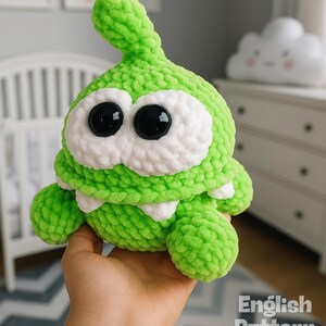 May include: A handmade green crochet plush toy with large black eyes and white eye surrounds. The toy has a toothy grin and small arms and legs. The text "PDF PATTERN" is at the top, and "English Pattern" is at the bottom.