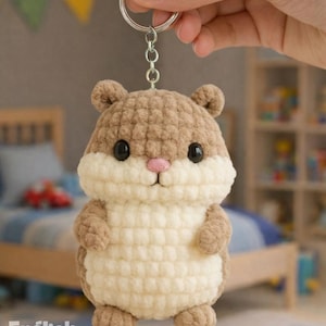 Puede incluir: Un llavero de hámster tejido a ganchillo en tonos marrón y crema, con ojos negros y nariz rosa, sujeto a un aro plateado. El texto en la imagen dice "PDF PATTERN" y "English Pattern".