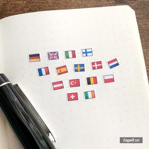 Può includere: Una collezione di piccoli adesivi rettangolari raffiguranti varie bandiere nazionali disposti su una pagina di un taccuino a puntini color crema, accompagnata da una penna stilografica nera con clip argentata posizionata sul lato sinistro.
