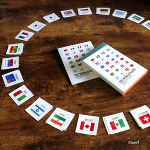Op de afbeelding: Een DIY World Flags memory game set met vlagkaarten gerangschikt in een cirkel. De speldoos toont verschillende vlaggen. Individuele vlagkaarten zijn verspreid rond de doos, waaronder vlaggen uit Australië, Italië en Canada. Het woord "Zagnet" is zichtbaar.