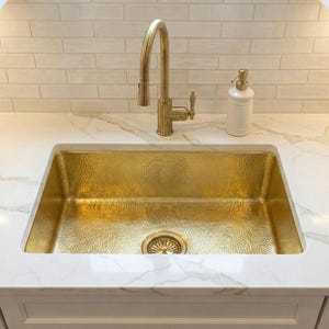 Op de afbeelding: Een gehamerde gouden gootsteen met bijpassende kraan en een witte keramische zeepdispenser. De gootsteen is geplaatst in een wit marmeren aanrechtblad met een witte bakstenen backsplash.